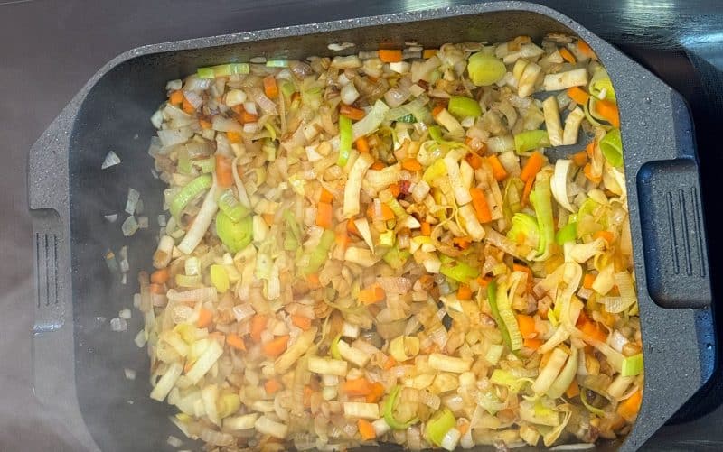 Ich gebe etwas zusätzliches Butterschmalz in den Bräter und füge die vorbereiteten Zwiebeln, Sellerie, Lauch, Möhren, Tomatenmark und Zucker hinzu. Alles lasse ich bei mittlerer Hitze langsam anbraten, bis es leicht braun wird.